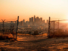 A photograph of a city from a distance, as seen through a ruined chain link fence. An orange haze sits over the city.