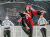 Three dancers on a large outdoor stage. Two male dancers, dressed in grey and black, flank a female dancer, dressed in black and red