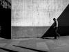 A black and white photo of a man walking down a sloping street in front of a wall transected by shadow