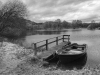 A black and white photo of a small boat tied to a dock at the edge of a wooded pond