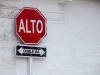 A stop sign displayed up against a white wall. Sign reads (in Spanish): Alto. Doble Via.