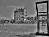 A black and white photograph of an empty town's square, with a chair that faces away from the viewer dominating the right side in the foreground