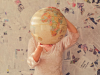 A woman stands in front of a wall covered in newspaper articles, holding up a tan colored globe that covers her face