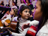 A young girl holding a guitar and surrounded by other children with instruments looks behind her