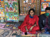 A woman clad in red sits with her husband (who is masked) on the ground. On the ground in front of them and mounted behind them on a wall of brightly colored paintings.