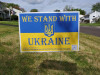 A photograph of a sign in a yard in a residential neighborhood. Text reads: We stand with Ukraine. 100% of all proceeds will go to the Ukrainian people.