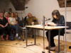 Friederike Mayröcker sits at a table reading with a small group sitting to the side