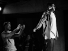 A black and white photograph of two actors on a stage. One stands with his arms thrust behind him, bathed in light, while the second stands behind, arms out front as if to catch him.