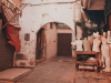 A photograph of the interior of city alley. Here, a collection of used mannequins gather outside of a stone archway