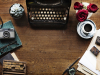 A top down shot of a writing desk with a manual typewriter, some plants, a cup of coffee, and implements associated with printmaking.