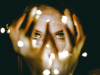 A photograph of a woman holding a string of lights in her hands, which are in front of her face. Only her eyes can be seen clearly through her splayed fingers