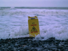 A "Caution: Wet Floor" sits on a beach and is enveloped by the tide