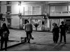 A black and white photo of a group of people, with its back to the viewer, walking toward a female figure, who stands in front of graffiti of a pair of angel's wings, painted on to the wall nearby