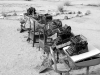 A black and white photograph of a line of typewriters on wooden desks on a beach. The desks and typewriters are covered with flotsam and sand.