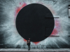 A photograph of a woman standing in front of the wall of an industrial looking building with a spray-painting of a black hole on the side of it