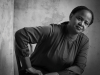 A black and white photo of Edwidge Danticat, seated but leaning forward slightly, with her hand on her leg