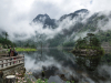 Spectators stand on a stone overlook that juts on to a wooded lake that sits at the foot of foggy mountains