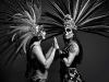 Two indigenous people, dressed in ceremonial makeup and dress, stand facing one another, palms touching