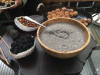 A spoon, partially submerged in a grain, rests on the edge of a wooden bowl, which itself is on a table with fruits and nuts