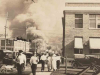 A sepia toned photograph. In the foreground, two white men pose with shotguns while others mill about. In the background, a smoke plume rises from a smoldering ruin