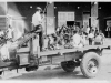 A black and white photograph. In the foreground is an open bed truck. In the bed, a man stands with a shotgun. An African-American man tends to the body of another, laid out on the bed.