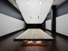 A photograph of an art installation. A long table sits beneath a long, curved light. The table is covered with carefully arranged parchment with writing on them.