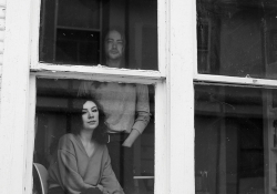 A black and white photo of a man, standing, and a woman, seated, looking through a window at the viewer