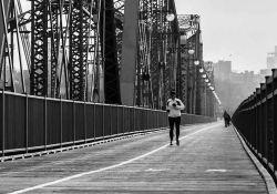 A black and white photograph of a person jogging toward the camera on a long suspension bridge