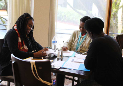 Three people sit around a table writing with a window illuminating the table