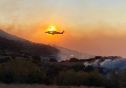 A helicopter hovers above a smoldering, mountainous landscape. The sun is positioned just such that the helicopter appears to be on fire.