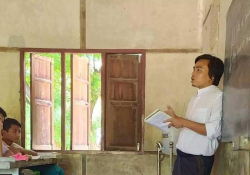 A teacher leans against the chalkboard as he speaks to the class. The first row of students can just be seen, looking up at him