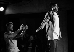 A black and white photograph of two actors on a stage. One stands with his arms thrust behind him, bathed in light, while the second stands behind, arms out front as if to catch him.