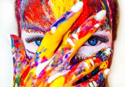 A close up on a female face streaked with a variety of different colored paints, her hand (also covered) is over her face but her fingers are split so her eyes are visible.