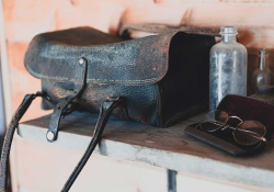 A weathered leather bag sits on a dresser with a dusty glass bottle and a pair of antique glasses sitting nearby