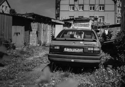 A black and white photograph of an old car parked behind a ramshackle apartment building in an overgrown alley