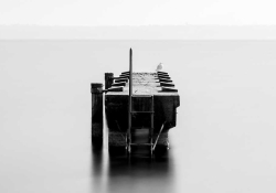 A black and white photo of a small boat dock surrounded by water, which stretches unbroken to the horizon