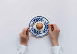 A photo looking down on a single egg on a fine piece of blue china, framed by two hands preparing to eat the egg