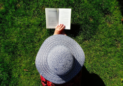 Person wearing a hat and laying in green grass reading a book.