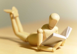 A close-up photograph of a small, wooden figure reading a tiny book.