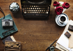 A top down shot of a writing desk with a manual typewriter, some plants, a cup of coffee, and implements associated with printmaking.