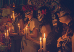 A group of people with their faces painted white like skulls hold candles before an altar