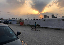 A young boy pauses on his bicycle on a gravel road with the sun either rising or setting behind a makeshift wall