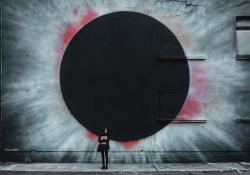 A photograph of a woman standing in front of the wall of an industrial looking building with a spray-painting of a black hole on the side of it