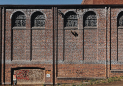 A small patch of graffiti mars the doorway of an imposing wall with tall windows up top. The wall has small text painted meticulously painted across its entire surface