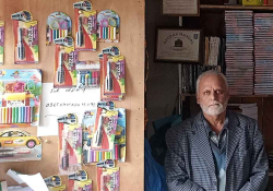 A photograph of an older man standing next to a wall with writing supplies on it