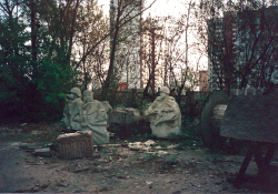 A collection of statues of resting soldiers, tucked into a small grove of trees with trash and industrial detritus strewn about